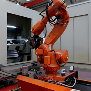 Orange robotarm som arbetar i en industriell miljö, placerad nära en maskin med en skärm synlig i bakgrunden.