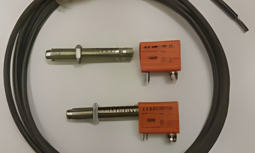 Två metallsonder med orange sensormoduler anslutna, var och en ansluten till en svart kabel, placeras på en vit yta. Enheterna ser redo ut för precisionsmätning eller testtillämpningar.