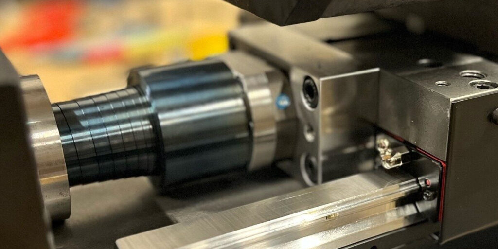 Närbild av industriella metallmaskiner med olika komponenter, inklusive en cylindrisk axel, metallytor och bälgar, i en verkstadsmiljö.