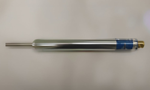 En metallcylinder med en mässingsfärgad kåpa och blå sektion i ena änden, som ligger horisontellt på en plan yta, verkar vara en del av precisionsenheter som används vid mätning eller tekniska uppgifter.