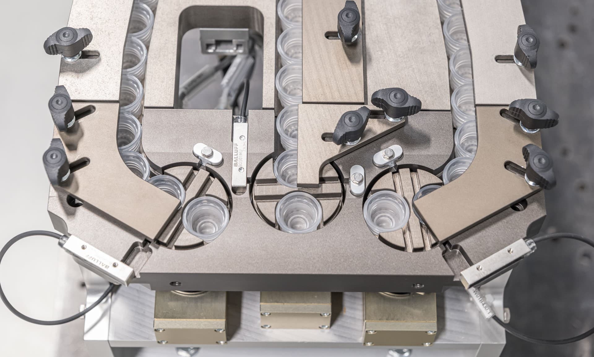 Industriell maskin med symmetriska metallarmar, rullar och slangar arrangerade runt en central arbetsyta, troligen för automatiserad montering eller hantering av material.